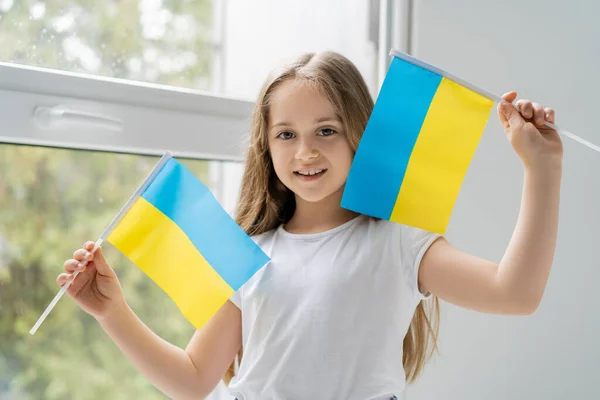 Ragazza con piccole bandiere ucraine sorridenti vicino alla finestra a casa — Foto stock