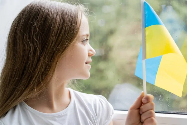 Niña sonriente con pequeña bandera ucraniana mirando a través de la ventana en casa - foto de stock