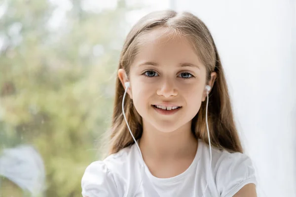 Alegre chica en auriculares con cable sonriendo a la cámara cerca de ventana borrosa - foto de stock