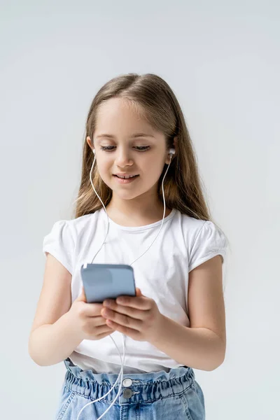 Sorridente ragazza in auricolare utilizzando il telefono cellulare isolato su grigio — Foto stock