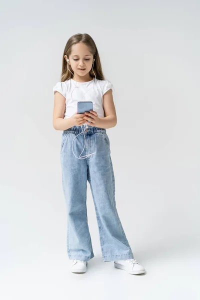 Vista a figura intera di ragazza in jeans e auricolari utilizzando smartphone su grigio — Foto stock