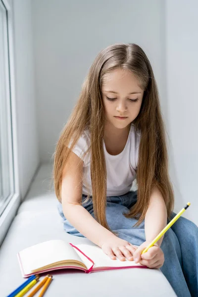 Mädchen mit langen Haaren sitzt auf Fensterbank und schreibt in Notizbuch — Stockfoto