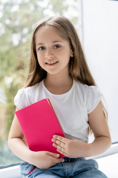 Fille heureuse avec les cheveux longs tenant livre de copie rose et souriant à la caméra — Photo de stock