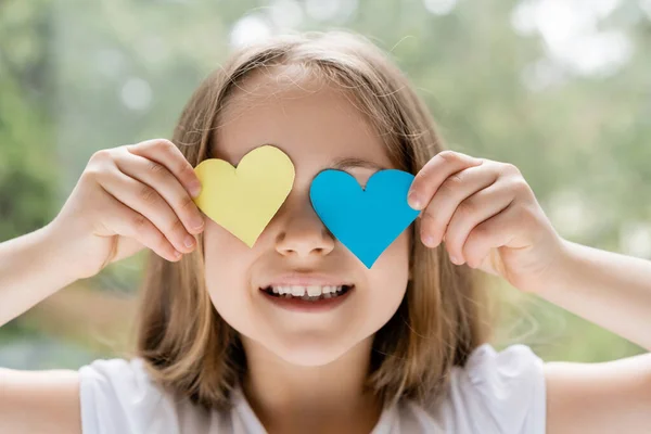 Menina ucraniana alegre obscurecendo rosto com corações de papel azul e amarelo — Fotografia de Stock