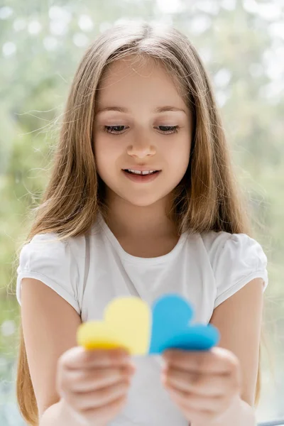 Lächelndes patriotisches Mädchen mit langen Haaren, das verschwommene blaue und gelbe Papierherzen hält — Stockfoto