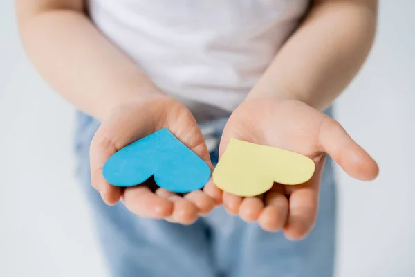 Vista recortada de un niño borroso sosteniendo corazones de papel azul y amarillo en palmas abiertas aisladas en gris - foto de stock