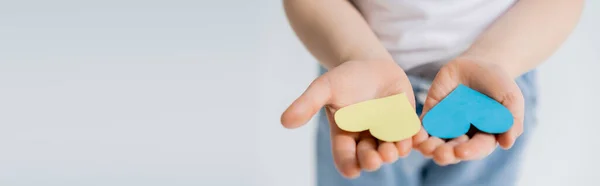 Vista parcial del niño con corazones de papel azul y amarillo en palmas abiertas aisladas en gris, pancarta - foto de stock