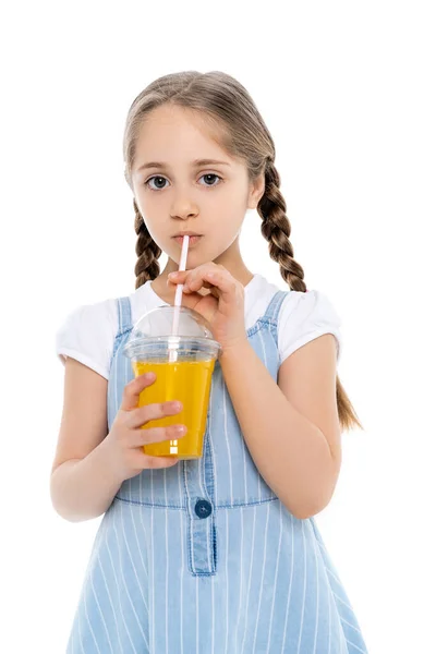 Mädchen in blauem Trägerkleid trinkt Orangensaft und blickt isoliert auf weiße Kamera — Stockfoto