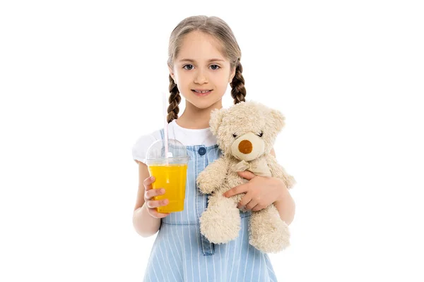 Fille heureuse avec du jus d'orange et un jouet doux regardant la caméra isolée sur blanc — Photo de stock