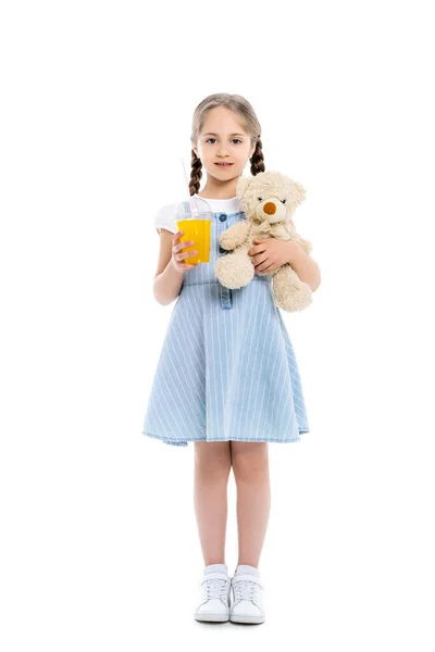 Full length view of girl in blue dress standing with orange juice and teddy bear on white — Stock Photo