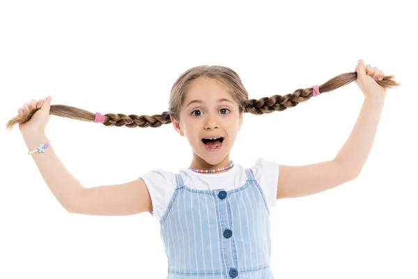 Excitada chica con la boca abierta sosteniendo trenzas y mirando a la cámara aislada en blanco - foto de stock