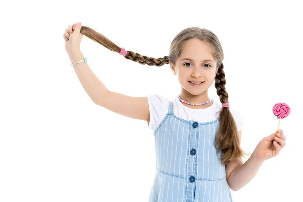 Chica sonriente con piruleta sosteniendo la trenza y mirando a la cámara aislada en blanco - foto de stock