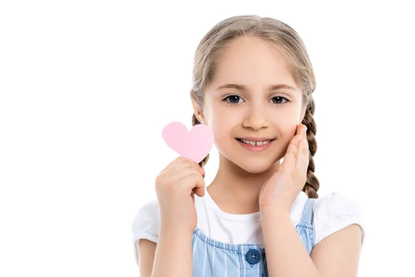 Compiaciuta ragazza toccare il viso mentre tiene il cuore di carta rosa isolato su bianco — Foto stock