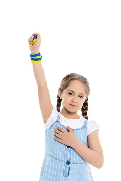 Patriotic ukrainian girl with blue and yellow ribbon touching heart while standing with raised hand isolated on white — Stock Photo