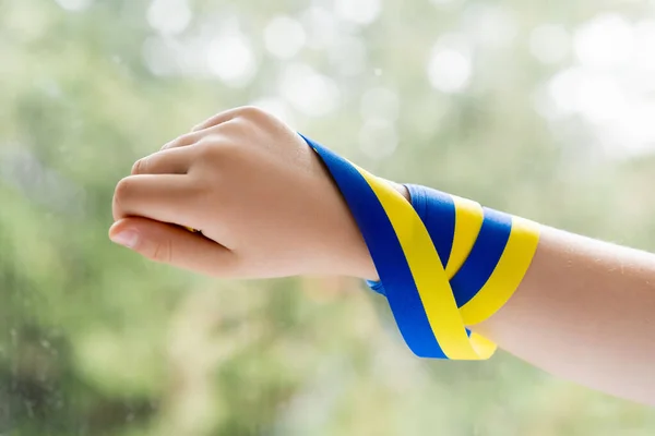 Vista recortada de niño con cinta azul y amarillo en la mano cerca de ventana borrosa - foto de stock