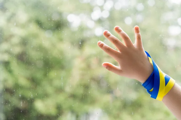 Abgeschnittene Ansicht eines Kindes mit blauem und gelbem Band, das Fenster mit Regentropfen berührt — Stockfoto