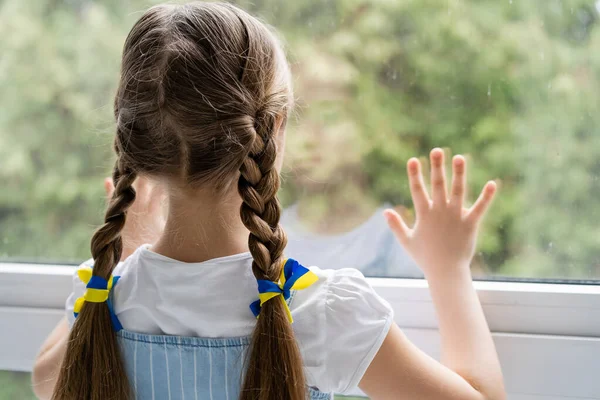 Back view of girl with blue and yellow ribbons on braids standing near window at home — Stock Photo