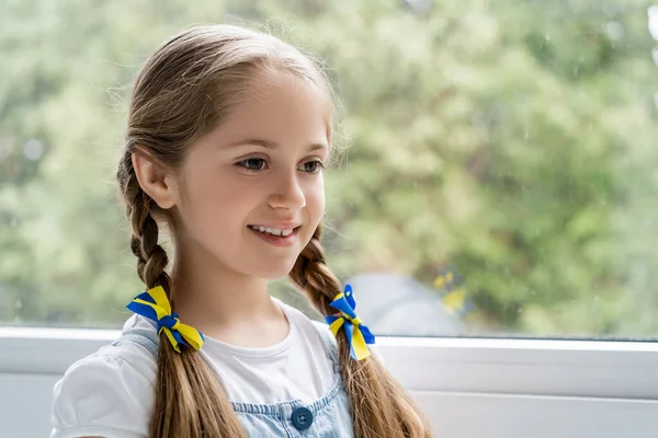 Ragazza patriottica con nastri blu e gialli sulle trecce sorridenti vicino alla finestra sfocata — Foto stock