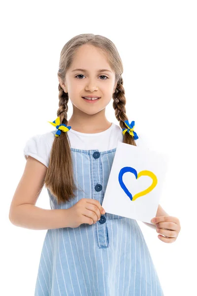 Niña con cintas patrióticas en trenzas que sostienen la tarjeta con corazón azul y amarillo aislado en blanco - foto de stock