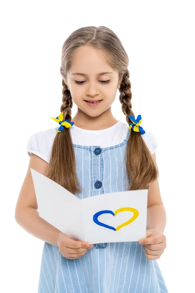 Girl with blue and yellow ribbons on braids reading card with heart sign isolated on white — Stock Photo