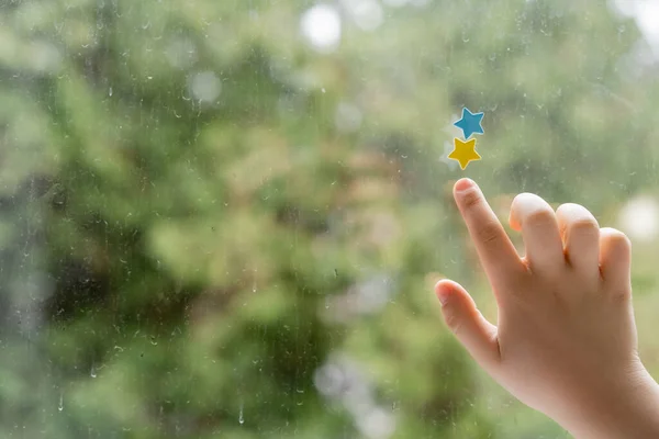 Vista ritagliata del bambino che punta le stelle blu e gialle sul vetro della finestra con gocce di pioggia — Foto stock