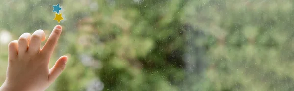 Vue recadrée de l'enfant touchant des étoiles bleues et jaunes sur la fenêtre avec des gouttes de pluie, bannière — Photo de stock