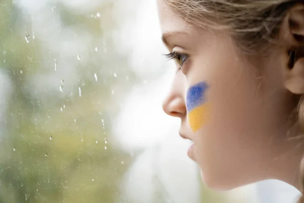 Primer plano perfil de chica con bandera ucraniana pintado en la cara cerca de vidrio ventana mojada - foto de stock