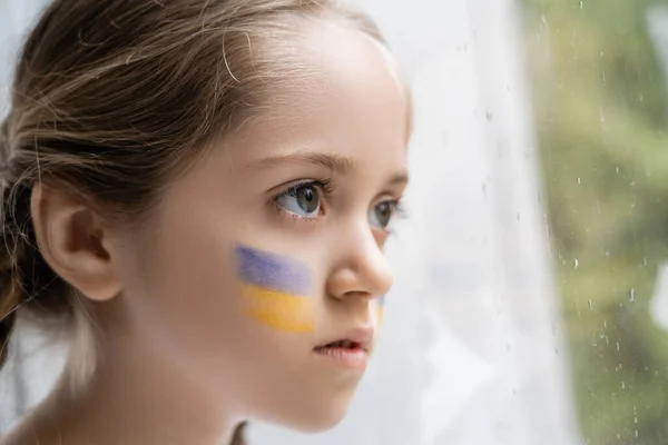 Portrait of girl with ukrainian flag on face looking away through window with raindrops — Stock Photo