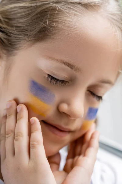 Primer plano retrato de chica con los ojos cerrados tocando la cara con banderas pintadas de Ucrania - foto de stock