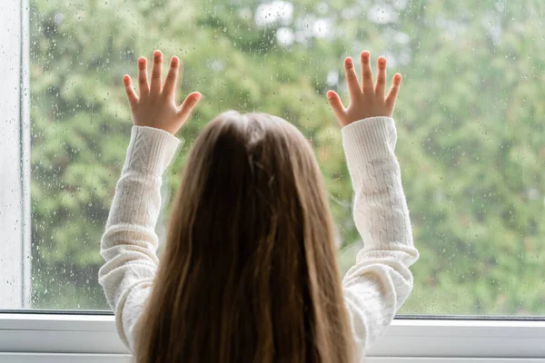 Back view of girl standing near window and touching glass with raindrops — Stock Photo