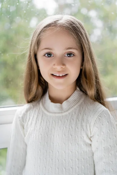 Glückliches Mädchen mit langen Haaren, das in die Kamera in der Nähe des verschwommenen Fensters blickt — Stockfoto