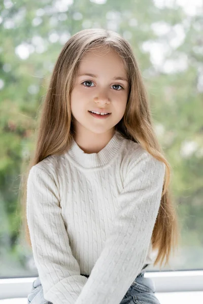 Chica positiva con el pelo largo sonriendo a la cámara cerca de ventana borrosa - foto de stock