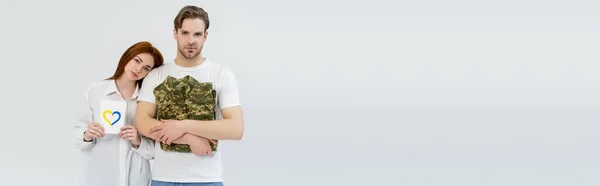 Woman holding card with blue and yellow heart near boyfriend with military uniform isolated on white, banner — Stock Photo