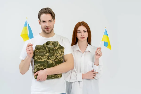 Pareja joven sosteniendo banderas ucranianas y uniforme militar aislado en blanco - foto de stock