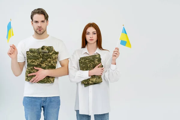 Pareja con banderas ucranianas y uniforme militar mirando a la cámara aislada en blanco - foto de stock