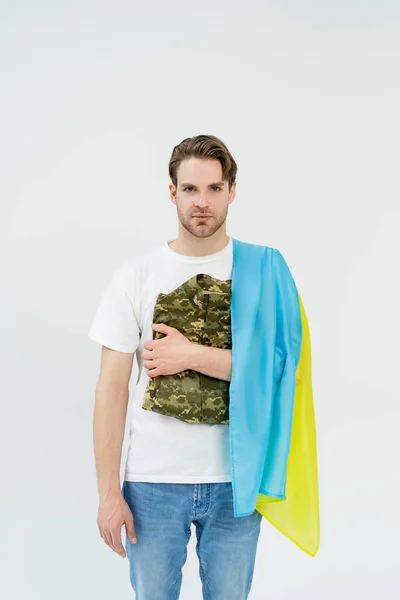 Joven con bandera ucraniana sosteniendo uniforme militar aislado en blanco - foto de stock