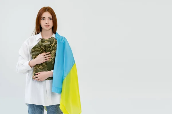 Mulher ruiva com bandeira ucraniana segurando uniforme militar isolado no branco — Fotografia de Stock