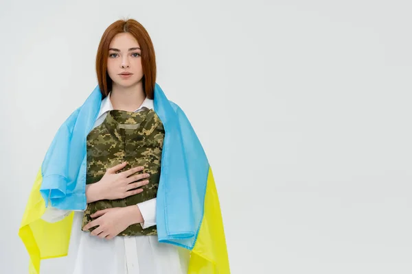 Mulher ruiva em bandeira ucraniana segurando uniforme militar isolado em branco — Fotografia de Stock