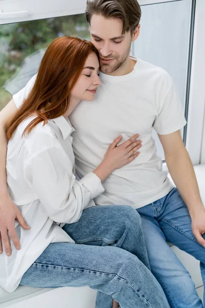 Hombre sonriente abrazando novia pelirroja en camisa en alféizar de ventana - foto de stock