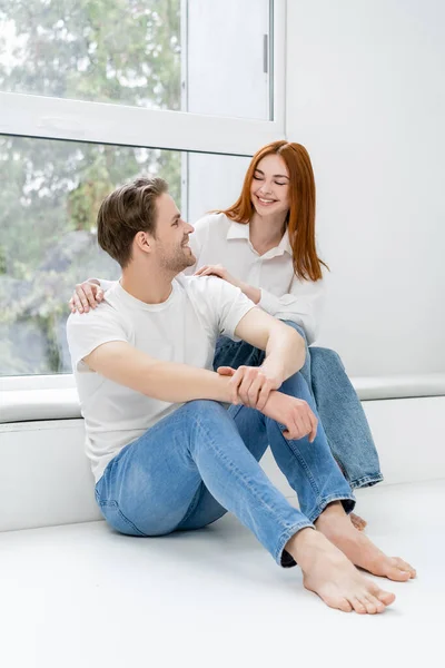 Pareja positiva mirándose el uno al otro cerca de la ventana en casa - foto de stock