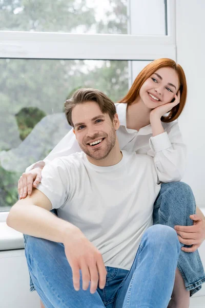 Sonriente hombre mirando a la cámara cerca de pelirroja novia en casa - foto de stock