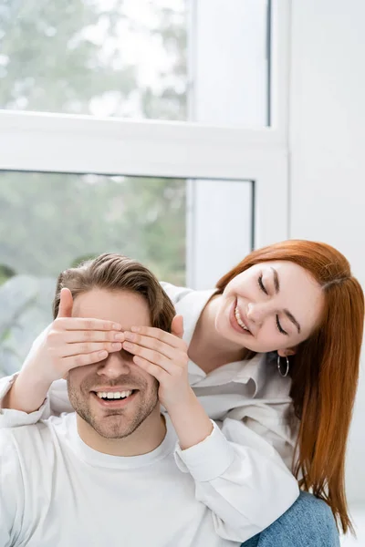 Mujer sonriente cubriendo los ojos del novio cerca de la ventana en casa - foto de stock