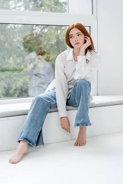 Redhead woman in jeans sitting on windowsill at home — Stock Photo