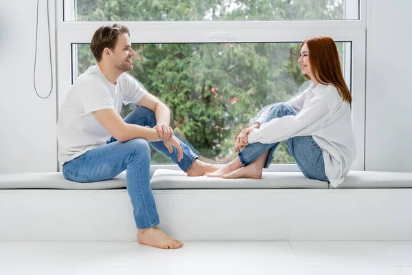 Vista lateral de pareja sonriente en jeans sentados en el alféizar de la ventana en casa - foto de stock