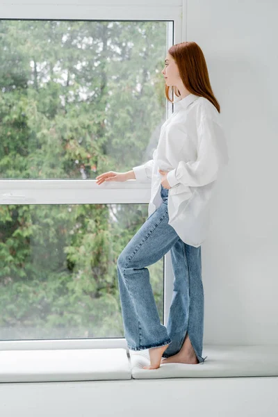 Vue latérale de la jeune femme en chemise et jeans debout près de la fenêtre — Photo de stock