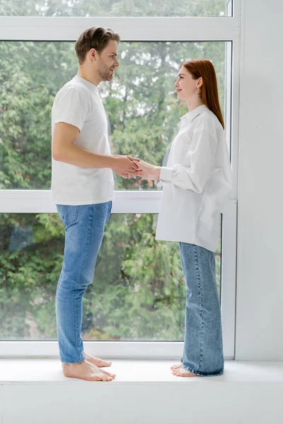 Vista lateral de pareja positiva descalza cogida de la mano en el alféizar de la ventana en casa - foto de stock