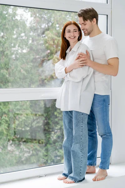 Uomo sorridente abbracciando rossa ragazza vicino alla finestra a casa — Foto stock