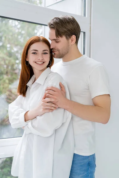 Joven abrazando alegre novia en camisa cerca de ventana en casa - foto de stock