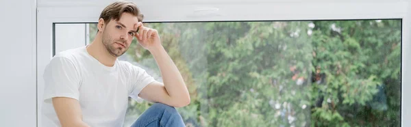 Jeune homme regardant la caméra près de la fenêtre à la maison, bannière — Photo de stock