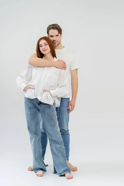 Full length of barefoot man hugging girlfriend and looking at camera on white background — Stock Photo